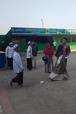 The video shows a gathering of people, mostly young males wearing white skullcaps and sarongs, in what appears to be an outdoor courtyard or walkway next to a building. The building has an orange and green facade with large windows. Some individuals are seated on the ground, while others are standing or walking. In the foreground, two individuals are walking away from the camera. In the background, a woman in a headscarf is visible. There are also some signs on the building, including one that reads "CANTEEN" and another with the number "8". The ground is paved with interlocking bricks. The overall atmosphere appears to be that of a school or religious institution.