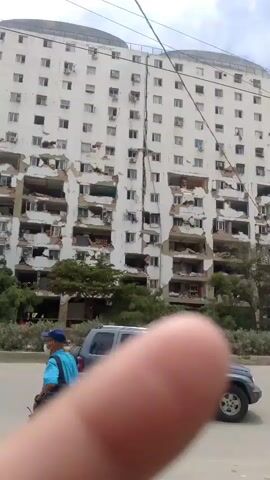 The video shows a partially collapsed apartment building. The facade of the building is white, and several balconies and sections of walls are missing, revealing the interior structure. Debris is visible on several floors. In the foreground, a person is walking past, and a car is parked on the side of the road. The scene appears to be outdoors, with trees and a cloudy sky in the background. The camera perspective is low, and a finger is in the foreground, partially obscuring the view.