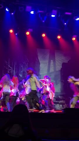 A low-angle full shot shows many dancers on a stage. Blue and red stage lights illuminate a stage with a background image of a city street and bridge in purple. About twenty dancers in white shirts and dark pants are moving in unison to their left and right. One dancer is held up by several dancers, while another group of dancers is holding another dancer. A person sits in the foreground with their back to the camera.