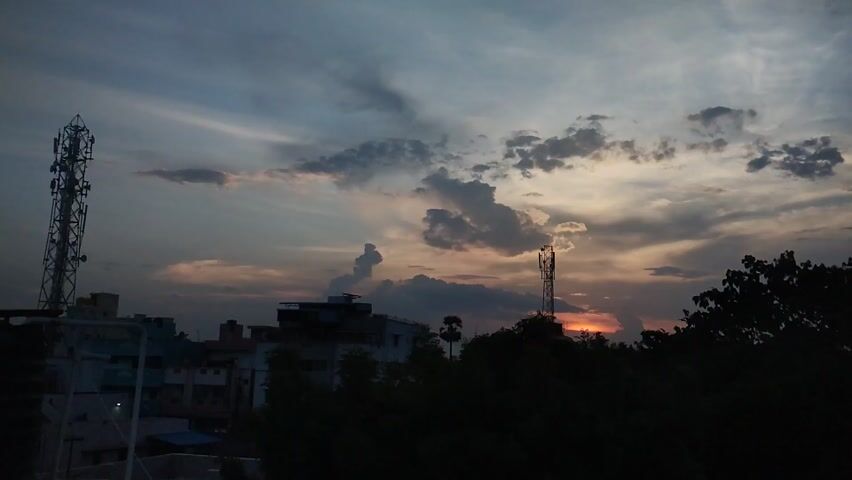 This video captures a sunset scene in Maduravoyal, India. The sky is filled with dramatic clouds, painted with hues of orange, pink, and gray, indicating either the setting or rising sun. Two tall cell towers are prominent features in the landscape, one on the left and another slightly to the right of center. In the foreground, dense, dark foliage obscures the ground level, suggesting a lush, possibly overgrown, area. Behind the trees and towers, the silhouettes of buildings are visible, hinting at an urban or semi-urban environment. The time of day is clearly sunset or dusk, given the warm, low-angled light and the darkening sky. The weather appears to be cloudy, with the clouds contributing to the picturesque and slightly dramatic atmosphere. No people are visible in the frame. A white chimney-like structure is visible in the bottom right of the frame, and a small portion of a sign with the text "PLUS" is visible on the far right. The overall mood is serene, with a touch of natural drama from the sky's colors and the stark industrial elements of the cell towers.