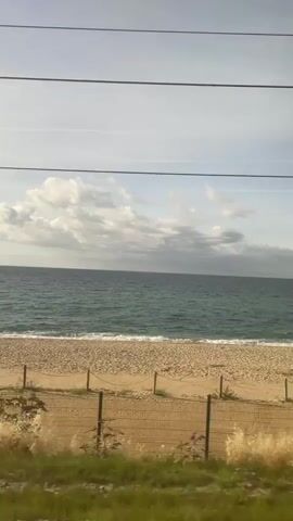 The video shows a view from a moving vehicle, likely a train, looking out towards a beach and the ocean. The sky is partly cloudy with a mix of white and gray clouds. The ocean is a deep blue-green color with gentle waves lapping at the shore. The beach is a light sandy color. In the foreground, there is a grassy area with some vegetation. A wire fence runs horizontally across the lower part of the frame, separating the grass from the beach area. As the video progresses, various structures and objects appear along the beach. Initially, there are some wooden posts and a wire fence. Then, a small wooden building with a metal roof comes into view. Further along the beach, there are construction materials, including piles of sand, orange cones, white bags, and what appears to be building supplies, suggesting ongoing work or development. There are also some small trees or bushes along the edge of the beach. The camera continues to move, showing different sections of the beach and the ocean.