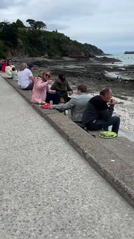 A medium shot shows a row of people sitting on a concrete wall by the sea on a cloudy day. Several adults and one child are seated, facing the ocean. In the foreground, two women and two men are eating seafood and drinking what appears to be sparkling wine. The woman closest to the camera is wearing a pink jacket and sunglasses, eating from a red platter. A man beside her, wearing a dark green jacket and a baseball cap, is also eating. Another man in a grey shirt is eating a similar dish. The beach in the background is rocky and exposed, indicating low tide, with some boats in the distance. The shoreline features cliffs covered in greenery.