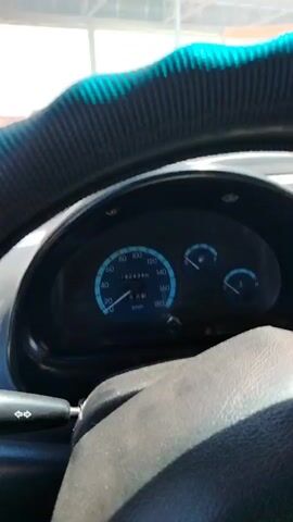The video shows the interior of a car. The dashboard gauges are visible, including a speedometer showing a speed of approximately 0 km/h. Outside the car, other vehicles are visible, and the scene appears to be a toll booth or checkpoint. One of the cars has a license plate that partially reads "242...4". The weather appears to be sunny. The perspective shifts from looking forward through the windshield to looking out the side window and into the side mirror. The side mirror shows reflections of other cars.