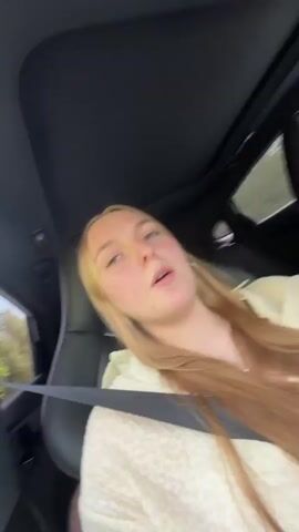 A young woman films herself in the passenger seat of a car, lip-syncing along with a song. She has long, strawberry blonde hair, fair skin, and a white shirt with a subtle animal print. She looks into the camera and gestures with her hands to the music. The driver, a young man with brown hair, is visible in the background, focused on the road. The car has a black interior and a sunroof. The surroundings seen through the window appear to be a rainy or overcast day.