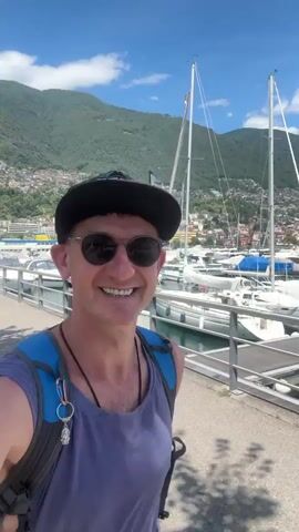 A smiling man wearing a purple tank top, sunglasses, and a baseball cap walks towards the camera, holding a cell phone to record a selfie video. He has a backpack on his shoulders, and a metal chain with a key hangs from his neck. Behind him, a marina is filled with docked sailboats and smaller vessels. Mountains covered in buildings and greenery rise in the background, under a bright blue sky with scattered clouds. The man is walking on a paved path next to a railing overlooking the water.