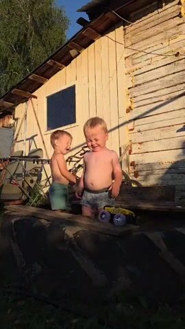 Two young boys, likely toddlers, are playing outside on a sunny day near a rustic wooden building. The taller boy, with blonde hair, is wearing light-colored patterned shorts and appears to be looking down at something on the ground. The shorter boy, also blonde, is wearing teal shorts and is holding a white toy. They are positioned on a large, dark tire, which serves as a platform. A yellow toy monster truck with purple wheels is visible on the tire near the taller boy. The building behind them is weathered and appears to be made of horizontal wooden planks. A window with a dark pane is visible on the building. In the background, a tree with green leaves and some metal structures, possibly part of a fence or equipment, can be seen. The lighting suggests it is late afternoon or early evening, with long shadows cast. The overall scene conveys a sense of simple, outdoor childhood play in a rural or semi-rural setting. The word "TORNADO" is visible on a bicycle part in the background.