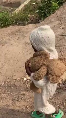 The video shows a small child wearing a hooded, white jumpsuit and green sandals, walking in a dusty outdoor environment. The child is carrying a brown teddy bear, hugging it close to their chest. The background includes trees, greenery, a partially constructed brick building, and some scattered debris on the ground. The child appears to be walking away from the camera, with the camera following their movement. The lighting suggests it is daytime.
