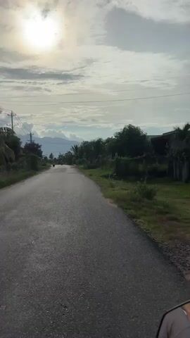 A first-person perspective video shows a paved road leading through a rural landscape. The sun is visible in the upper left corner, partially obscured by clouds. The road is lined with green grass, trees, and some buildings. Two motorcycles are seen in the distance on the road. In the background, mountains are visible under a cloudy sky. A bird is briefly seen flying in the sky. The video appears to be taken from a moving vehicle, likely a motorcycle, as indicated by the lower right corner of the frame which shows part of a hand and a vehicle handle.