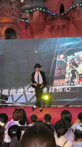 A male performer dressed as Michael Jackson, wearing a black suit, white shirt, black tie, and fedora hat, performs on a stage in front of a large screen. He is facing the audience and the screen, which displays images of a cage and people. The stage is covered with a red carpet, and several spotlights are positioned on the stage. Behind the stage, a brick building with decorative lights is visible. An audience of people, mostly facing away from the camera, is seated in front of the stage, many of whom are holding up phones to record the performance. The performer is in mid-dance, with his arms extended and legs bent, mimicking Michael Jackson's iconic dance moves. The video captures the performer's movements and the reactions of the audience.