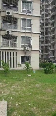 The video shows a partial view of a building's exterior, focusing on a section where two individuals are working on scaffolding. The building appears to be multi-story, with windows and air conditioning units visible. The scaffolding is constructed from bamboo poles and is attached to the side of the building, allowing access to an upper window. Both individuals are positioned on the scaffolding, and appear to be engaged in painting or a similar maintenance task, as they are holding buckets. The ground level features a grassy area with some small plants and trees. In the background, another, taller building is visible. A white van is also partially visible to the right of the frame. The overall scene suggests an outdoor construction or maintenance activity.