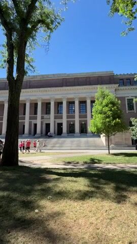 The video shows a sunny day with a clear blue sky. In the foreground, a large tree with green leaves casts a shadow on a grassy area. Several people are visible in the middle ground, standing on a path or lawn in front of a large building. The building has a classical architectural style with a long facade, a portico with white columns, and a brown roof. Some people are standing in small groups, while others are walking. The overall scene appears to be a campus or public space on a pleasant day.