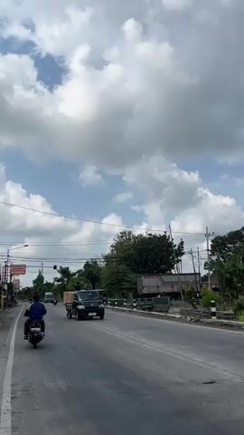 The video shows a road with some traffic. In the foreground, a black pickup truck is moving towards the right. It has a wooden crate in the bed and two men sitting on the side of the truck bed. Behind this truck, another black pickup truck is visible. Further down the road, a motorcycle is moving towards the left. The sky is partly cloudy with white clouds against a blue sky. Trees and buildings are visible on the sides of the road. The road appears to be asphalt.