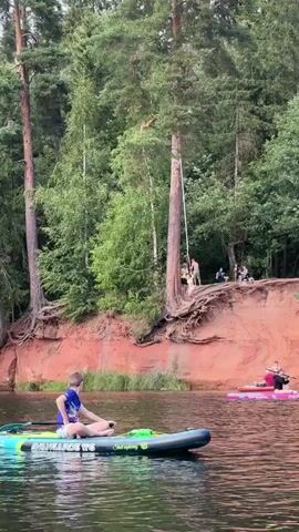 A young boy sits on a paddleboard in a lake. He wears a blue t-shirt and shorts. In the background, people are gathered on a sandy cliff overlooking the water. A person is captured mid-air, jumping from the cliff into the lake, creating a splash. Another person on a paddleboard is visible in the distance. The environment is a wooded area with tall trees and a red, sandy cliff face.