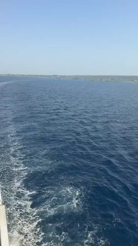 The video shows a view from the side of a moving vessel. The water is a deep blue with white wakes trailing behind the boat. In the distance, a coastline is visible with some vegetation and buildings. The sky is clear and pale blue, gradually becoming brighter and hazier towards the horizon where the sun is shining, casting a glare on the water. A metal railing is visible in the lower right corner of the frame in the final seconds.