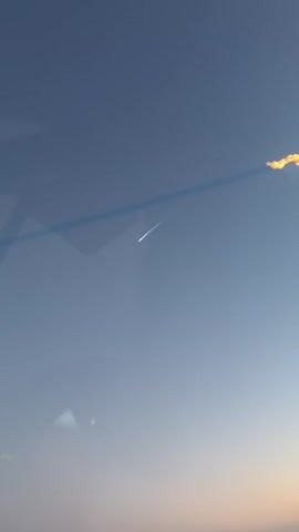 A video captures a clear blue sky with a faint line of orange near the bottom horizon. A thin, white contrail stretches diagonally across the sky from the upper right to the lower left. The contrail appears to be from a high-flying aircraft or possibly a shooting star, though it is too indistinct to be certain. There are some subtle reflections on the surface from which the video was taken, appearing as faint geometric shapes and hazy colorful spots. The overall impression is one of a serene, clear sky with a visible atmospheric phenomenon.