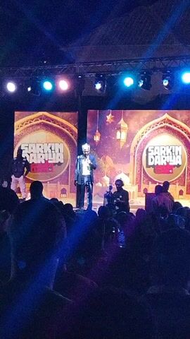 The video shows a stage performance in front of an audience. A man in a suit is speaking into a microphone on the stage, illuminated by stage lights. Behind him are two large screens displaying the text "Sarkin Dariya" and an illustration suggesting an Islamic theme with lanterns and a mosque. Another person stands near the speaker, potentially a security guard. A cameraman is also present on stage, filming the event. The audience is seated in the foreground, with their backs to the camera, observing the performance. There are more lights and scaffolding above the stage.