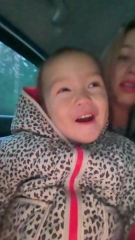The video shows a young child sitting in what appears to be the back seat of a car, looking directly at the camera. The child is wearing a hooded jacket with a black and white animal print and a pink zipper. They are smiling and appear to be happy. A woman with blonde hair and red lipstick is partially visible behind the child, also in the car. The interior of the car is visible, including the ceiling and window. The video seems to be taken at dusk or in a dimly lit environment.