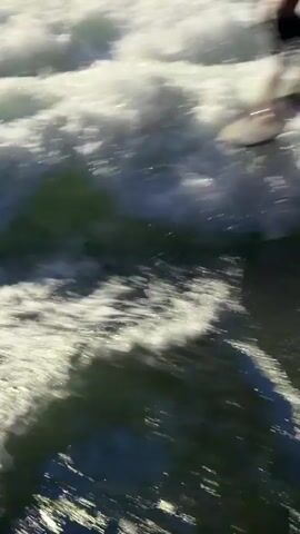 A surfer rides a wave in the river. The surfer is shirtless and wearing dark shorts. The surfer is in the middle of the wave, with white water spraying around them. There are several people on the bank of the river, watching the surfer. The people on the bank are holding surfboards. The river is flowing rapidly, and the waves are choppy. The sun is shining, and there are shadows on the water.