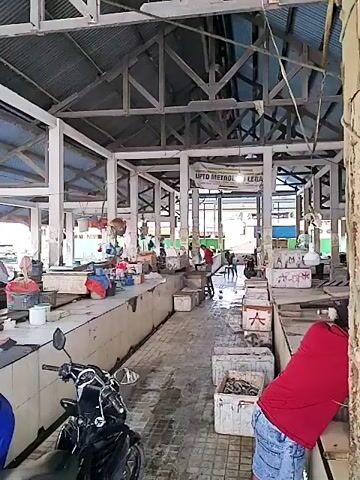 The video shows an indoor market, possibly a fish market, with several stalls. The structure appears to be made of wood and corrugated metal. There are a few people present, some of whom appear to be vendors. In the foreground, a motorcycle is parked. Some of the stalls have a variety of items displayed, including what look like fish in crates and buckets. The floor is tiled and appears to be wet in places. There is a sign with some text that is partially visible. The lighting suggests it is daytime.