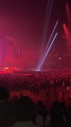 A wide-shot vertical video shows a large crowd at a music concert venue. The venue is dark, bathed in intense red light from stage lights. Two prominent beams of white light shine down from the ceiling towards the stage, illuminating a figure in the distance. The crowd is dense, with many people holding up phones, presumably to record the performance. Some individuals are more clearly visible in the foreground, while the crowd further back appears as a mass of silhouettes. Speakers and stage rigging are visible in the background. Some orange lights appear further to the right near the top of the frame.