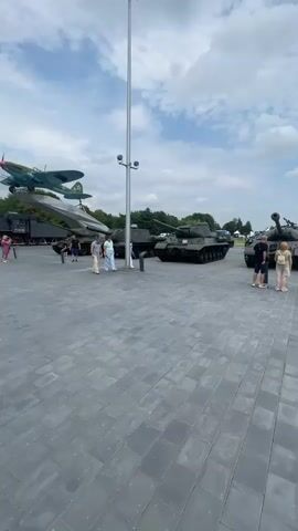 The video shows an outdoor exhibition of military vehicles, including several tanks and an airplane displayed on a raised platform. The scene is set in a public area with a paved ground made of gray rectangular blocks. People are seen walking around the exhibition, observing the vehicles. The weather appears to be cloudy with patches of blue sky, suggesting it might be daytime, possibly late morning or afternoon. The overall atmosphere seems casual and educational, as visitors are exploring the historical display. No visible text is present in the video. The setting suggests a museum or memorial dedicated to military history.