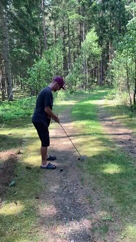A man is depicted attempting to hit a golf ball with a club in a sunlit forest setting, likely in Vessigebro, Sweden. He wears a dark t-shirt, dark shorts, a red/purple baseball cap, and flip-flops. He holds a golf club, possibly a putter or a wedge, and positions himself over a golf ball on a dirt and grass path. As he takes a powerful swing, he loses his balance, spins around, and almost falls over, exclaiming "Oh!" in surprise or amusement. The scene is bright, indicating daytime, with tall pine trees surrounding the path and casting shadows. The ground is a mix of dirt, grass, and moss, typical of a forest clearing or trail. No visible text is present.