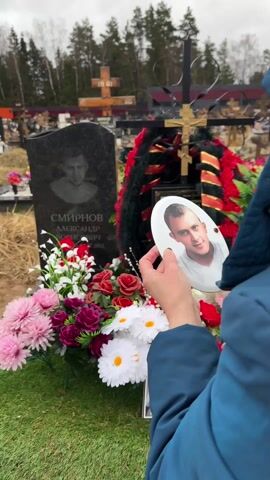 A person is holding a photograph of a young man in front of a gravestone. The gravestone is engraved with a portrait of the same man and the name "СМИРНОВ АЛЕКСАНДР БОРИСОВИЧ" (Smirnov Alexander Borisovich). The gravestone is adorned with colorful artificial flowers. Behind the gravestone, there's a black and gold cross decorated with a wreath. In the background, there is a cemetery landscape with other graves and trees. The person holding the photo is wearing a blue jacket. The scene is somber and appears to be a memorial visit to a gravesite.