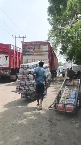 In Fagge A, Nigeria, on a bright, sunny day, a man is seen pushing a cart heavily laden with cases of Coca-Cola bottles. The cart is stacked high with these cases, creating a precarious but impressive tower of product. The man, wearing a blue t-shirt and dark shorts, is focused on maneuvering the cart.

Behind the cart, a large red truck is parked, its trailer also displaying a prominent Coca-Cola advertisement with the slogan "Whichever way you like it, there's a Coke for you." This truck appears to be the source of the goods being transported. Another red truck is visible to the left of the primary truck.

The setting is an open, dusty area, possibly a distribution center or market. The ground is uneven and dirt-covered. Trees with green foliage line the right side of the frame, providing some shade. In the background, other people can be seen, some walking and others possibly working, suggesting a busy commercial environment. A second, smaller cart filled with bottled water is also visible in the foreground on the right.

The overall impression is one of active commerce and product distribution, with the abundant Coca-Cola products and the transportation vehicles highlighting the theme. The bright sunlight suggests it is midday or afternoon.