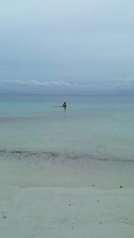 The video begins with a wide shot of a calm ocean and a sandy beach. The water is a clear turquoise color near the shore, gradually deepening to a bluish hue further out. The sky is overcast with a few wispy clouds. In the distance, a person appears to be wading in the water. As the video progresses, another person walks into the water from the left side of the frame. The camera then shifts to a closer view of the shoreline where several people, including children, are interacting in the shallow water. A red boat with people on board is anchored nearby. A person in the foreground bends down to pick up something from the sand. The scene depicts a relaxed day at the beach with people enjoying the water and the scenery.