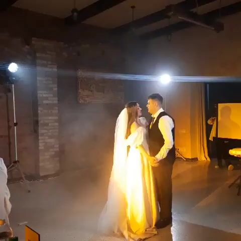 A wedding reception is shown, with a newly married couple dancing. The bride is wearing a white wedding dress and veil, and the groom is in a black vest and white shirt. They are slow dancing in the center of the room, surrounded by a light fog effect. Spotlights illuminate the couple, casting golden light on them. The background of the room features exposed brick walls and a wooden ceiling with exposed beams. There are lights on stands and a projector screen visible in the background. The overall atmosphere is romantic and celebratory.