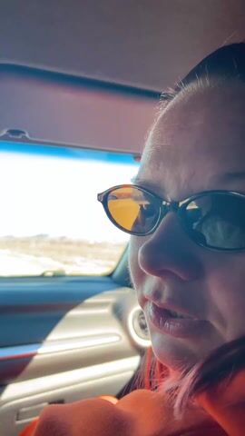 From inside the cabin of a car, a woman in an orange sweater wearing sunglasses is seen in the passenger seat. Her face is close to the camera as she speaks. She turns her head to look out the front windshield as the car travels on a highway. The landscape outside the car includes trees, fields and a bright, clear sky. Other vehicles can be seen on the road. The interior of the car is visible, including the dashboard, rearview mirror and sun visor.