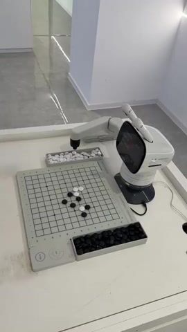 The video shows a white robotic arm that is playing the game of Go against a human player. The game board is on a white table, with white and black game pieces on the board and in trays on either side of the board. The robot arm has a screen that displays information, including the remaining time. The background is a hallway with white walls. A person is briefly seen walking past in the background, carrying a water bottle and a phone.