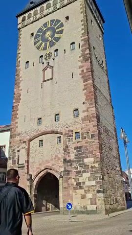 The video shows a tall stone tower with a large clock face. A man in a black shirt and shorts walks towards the tower, entering a large arched opening at its base. Other people are seen walking and cycling in the background on a cobblestone street. The sky is clear and blue, indicating a sunny day.