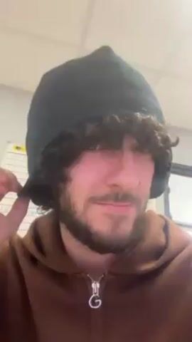 A young adult male removes a dark beanie from his head, revealing thick curly hair. He is wearing a brown hoodie with a zipper and a logo on the front. He looks directly at the camera and smiles slightly. The background appears to be an office or workspace with a whiteboard or calendar visible. The lighting is bright and appears to be artificial.