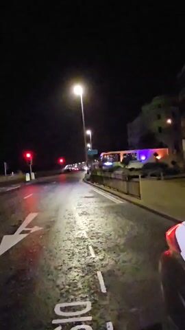 A point-of-view night video shot from a moving vehicle on an asphalt road. The road is wet, and street lights illuminate it. Road markings, including directional arrows and the word "DIS" are visible. In the distance, red traffic lights are illuminated. On the right, a building with blue and red lights can be seen beyond a railing. The sky is dark.
