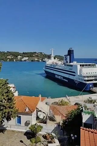 The video shows a large ferry boat docked at a harbor. The ferry is blue and white with "SEA JETS FERRIES" written on its side. There are several cars and people on the dock near the ferry. In the foreground, there are buildings with red tile roofs. The water is blue and clear, and the sky is a bright blue. The scene appears to be a sunny day in a coastal town.