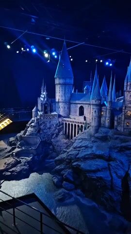 The video shows a detailed model of Hogwarts castle, illuminated by blue spotlights. The castle sits atop a rocky landscape, surrounded by simulated water. The camera slowly pans from left to right, providing a view of the castle’s various towers, architectural details, and the surrounding terrain. A railing is visible in the lower-left corner of the frame, suggesting the viewer is observing the model from a designated area. The background is dark, drawing focus to the illuminated castle.