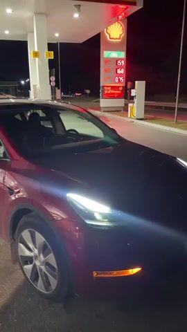 A red Tesla, with its headlights on, is parked at a Shell gas station at night. The gas pump display shows prices for different fuel types. The gas station has a white roof and pillars. The road and trees are visible in the background.