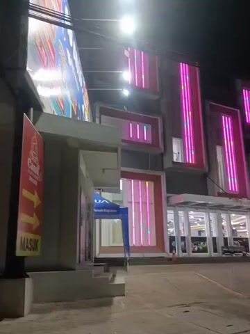 The video shows the exterior of a modern building at night. The building has multiple floors and features prominent pink and white vertical LED lights that illuminate the facade. The lights are arranged in rectangular patterns on different sections of the building. In front of the building, there is a paved area that appears to be a parking lot or walkway. A few people are visible walking near the entrance of the building, which is glass-fronted and appears to be a commercial establishment. There are also some flags and banners, including what appears to be an Indonesian flag. To the left of the building, a large, illuminated billboard with some text and images is visible. There is also a smaller structure with a blue awning and some signage. The overall impression is of a well-lit, contemporary building at dusk or night.