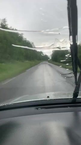 The video is taken from inside a vehicle looking out through the windshield. It is raining, as evidenced by raindrops on the windshield and the wet road ahead. The road appears to be a two-lane asphalt road with significant damage, including numerous potholes and cracks. The road is lined with trees and vegetation on both sides, suggesting a rural or wilderness setting. The sky is overcast and gray, indicating a cloudy or stormy day. The vehicle appears to be moving forward along the road. There are no people visible in the frame.