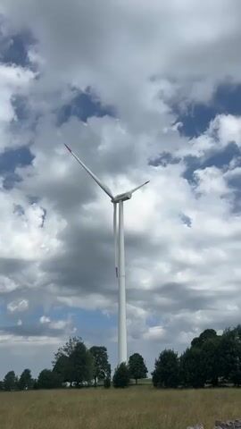 A vertical, low-angle shot captures a tall, white wind turbine against a cloudy sky. The turbine's blades are white with red tips and are in motion, indicating it is active. The turbine stands in a field with dry, yellow-brown grass in the foreground and a line of green trees in the middle ground. The sky is a mix of white and gray clouds, with patches of blue visible. The overall impression is one of a windy, overcast day in a rural, open landscape.