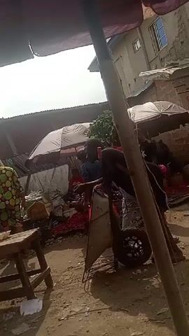 The video shows an outdoor marketplace with a variety of people milling about. There are several vendors with stalls set up under umbrellas, displaying various goods. Some people are carrying items, like a person carrying a bundle of green leaves on their head, and a man wheeling a small cart. Many people stand and talk in small groups near tables and displays. The ground appears to be dirt or packed earth. Some buildings can be seen in the background behind the stalls.