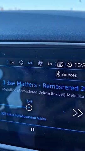 The media file shows a close-up of a car's infotainment screen displaying music playback information. The screen is dark blue with bright blue text and icons.

**Subjects:** The primary subject is the car's infotainment system screen, displaying song details. There are no people or animals visible.

**Objects:** The objects include the car's dashboard, the infotainment screen itself, and various icons on the screen. These icons represent climate control (Lo, AC, fan speed), time, Bluetooth, and playback controls (play/pause, next track).

**Scene/Setting:** The scene is the interior of a car. The dashboard surface is visible at the top of the frame, textured and dark in color. The lighting suggests it is daytime, possibly with sunlight contributing to the glare on the dashboard, although the exact time of day is not explicitly stated.

**Activity/Event:** The main activity is music playback. The screen shows the song title "Nothing Else Matters" by Metallica from the "Remastered Deluxe Box Set." The current playback time is displayed as "3:48" and the progress bar indicates it's nearing the end of the song. There is also text indicating the audio source is "S25 Ultra пользователя Nikita."

**Notable Details:** The song title and artist are clearly visible, indicating the genre is rock. The "S25 Ultra" likely refers to a Samsung Galaxy S25 Ultra phone, suggesting the music is being played via Bluetooth from a user named Nikita. The climate control settings are also visible, showing "Lo" for temperature and the AC is on, with a fan speed icon.

**Visible Text:**
* "Lo" (repeated twice)
* "A/C"
* "Nothing Else Matters"
* "Metallica (Remastered Deluxe Box Set)"
* "3:48"
* "S25 Ultra пользователя Nikita"
* "Sources"