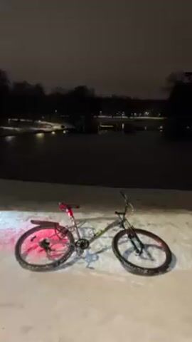 A bicycle stands stationary on a snow-covered path. A faint red light illuminates the rear wheel and the ground behind it. In the background, a dark, frozen lake is partially illuminated by distant city lights. Trees line the distant edge of the lake. The sky is dark and overcast. The scene appears to be at night in a park or similar outdoor location.