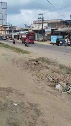 The video captures a daytime street scene in what appears to be a small town or rural commercial area in Manetunong, Indonesia, under an overcast sky.

In the foreground, there is an unpaved, dusty dirt path or shoulder strewn with debris, including discarded wood pieces and other refuse. A small concrete culvert or drain is also visible. The main focus is a paved two-lane road where traffic is moving from left to right.

The primary subject is a bright red truck, specifically a Mitsubishi Fuso, carrying numerous red gas cylinders in an open metal cage at the back. Text on the truck reads "PT. NONGAL SENTRAL GAS" and "Tabung Gas." Following closely behind the red truck is a smaller black delivery truck, possibly a Mitsubishi L300 or similar model, with a blue and white logo on its side, resembling bottled water branding (possibly "Aqua").

Other subjects include a person walking on the dirt path on the left side of the road, and a person riding a motorcycle further down the street behind the trucks. Several other motorcycles are parked along the roadside in the background.

The background features a row of two-story buildings, housing various shops and businesses, indicated by colorful signs. One visible sign on a building reads "MONAS." Utility poles with numerous overhead wires run parallel to the street, suggesting an established infrastructure. The overall impression is of a busy, yet somewhat unkempt, local commercial street during the day.