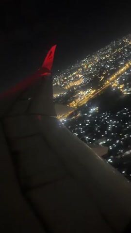 The video presents a nighttime view from an airplane window, looking down at the city lights of Addis Ababa, Ethiopia. The wing of the aircraft is visible in the foreground, partially obscuring the lower left of the frame. Below, a vast expanse of city lights forms a dense, glowing network of bright dots and lines, illuminating the dark landscape. The plane appears to be flying over or in the process of taking off or landing, offering a sweeping perspective of the illuminated urban sprawl against the dark night sky. The scene is clear, with no visible clouds.