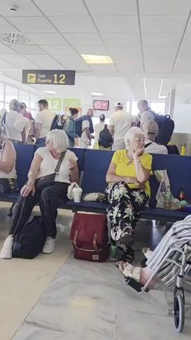 The video shows an airport departure lounge where several people, mostly older adults, are seated on blue benches. Many are looking at their phones or eating. A sign above indicates "Gate Puerta 12". The environment appears to be a typical airport waiting area with tiled flooring and fluorescent lighting. People are dressed casually in summery clothing, and several have bags and backpacks with them. The overall atmosphere suggests a waiting period before a flight.