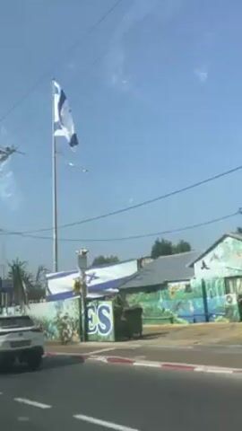 The video shows a view from inside a car driving along a road. On the right side of the road, an Israeli flag flies on a tall pole. The flag is tattered and flapping in the wind. Behind the flag is a building with a large Israeli flag painted on its side. There is a white Lexus car parked on the side of the road. The sky is blue and clear.