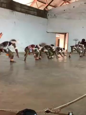 A group of barefoot men perform a traditional dance in a large, sparsely decorated room. They are dressed in white tank tops, printed green and gold patterned pants, and white headbands with black and red accents. The dancers start crouched low to the ground, moving their hands on the floor, then rise to a standing position, maintaining a synchronized rhythm. A woman, also barefoot, stands to the side, seemingly directing the dancers. She wears a patterned top, a short skirt with fringe, and colorful arm bands. The room has white walls, a concrete floor, and a corrugated metal roof. A doorway is visible in the background. The sound of drums is prominent.