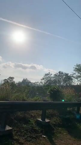 The video is a vertical shot of a rural landscape bathed in bright sunlight. In the foreground, a dark pole stands vertically on the left side of the frame, with a wire extending from its top across the sky. To the left of the pole, tall, wispy grasses are visible. The middle ground features a sloping area with young trees, some of which appear to be rubber trees, scattered across the terrain. The ground is covered in dry grass and some green vegetation. In the background, a line of trees forms a dense, dark silhouette against the bright sky. The sky itself is blue with scattered white clouds and the sun is prominently visible, casting a strong glare. A black metal railing or barrier is present in the lower portion of the frame, extending across the width of the image. The overall impression is of an outdoor scene, possibly taken at sunrise or sunset, with a natural, slightly unkempt environment.